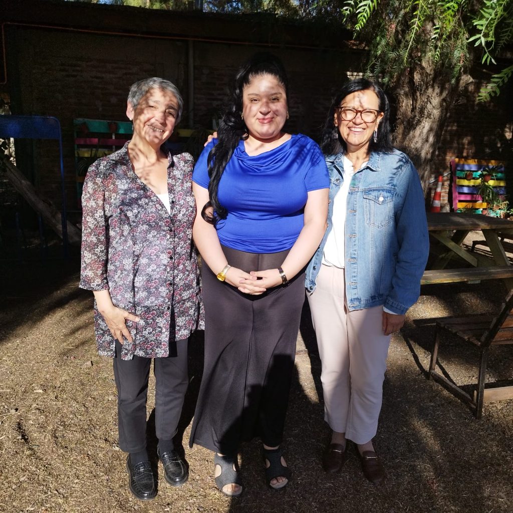 Tres mujeres de diferentes edades, sonriendo y de pie juntas en un patio comunitario en Chile, posan para la cámara, representando el espíritu del voluntariado en Chile.