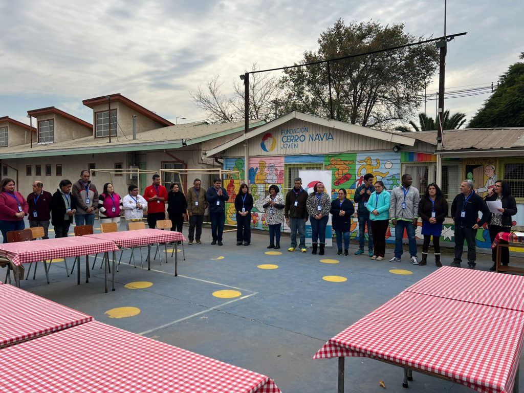 Actividad al aire libre de Bienvenida en Los Lagos. Un grupo de adultos, varios con credenciales, están de pie formando un semicírculo en un patio, frente a un edificio con un colorido mural de la Fundación Cerro Navia Joven.