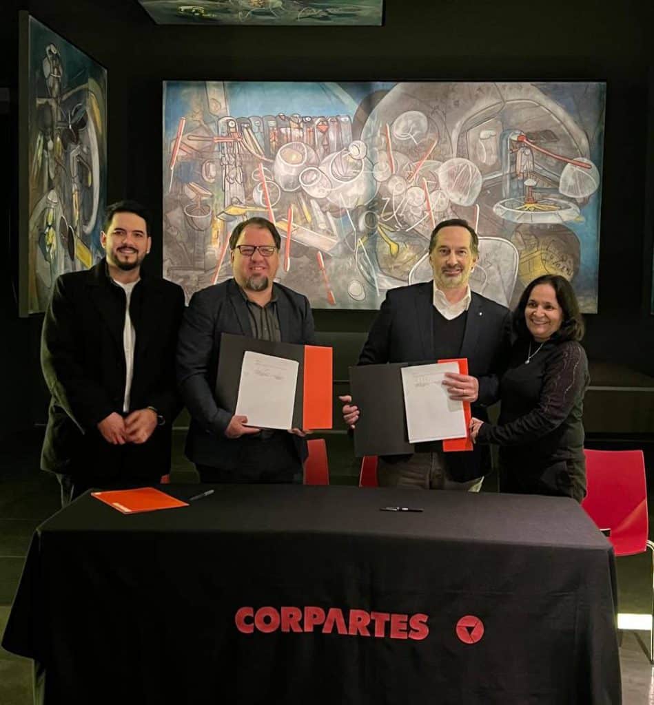 Fotografía de cuatro personas (tres hombres y una mujer) sonriendo a la cámara detrás de una mesa cubierta con un mantel negro que lleva el logotipo rojo de CORPARTES. Los dos hombres en el centro sostienen documentos firmados en carpetas formales, celebrando el momento en que renovamos convenio con CorpArtes.