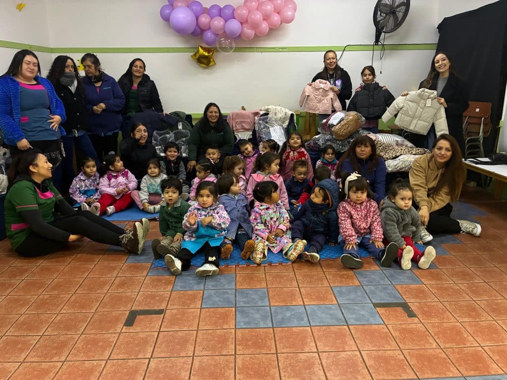 Un gesto de solidaridad: Una fotografía grupal en un espacio comunitario donde varias mujeres adultas y un grupo de niños pequeños están reunidos. Las mujeres, algunas de pie al fondo y otras sentadas, sonríen mientras sostienen y muestran pilas de ropa infantil nueva (como abrigos y chaquetas). Los niños están sentados juntos en primer plano sobre una manta azul extendida en el suelo de baldosas, mirando en varias direcciones. El fondo de la habitación tiene paredes blancas y verdes y está decorado con un racimo de globos de colores rosa, morado y dorado. Esta imagen captura un momento de apoyo comunitario a través de la donación de ropa para los niños.