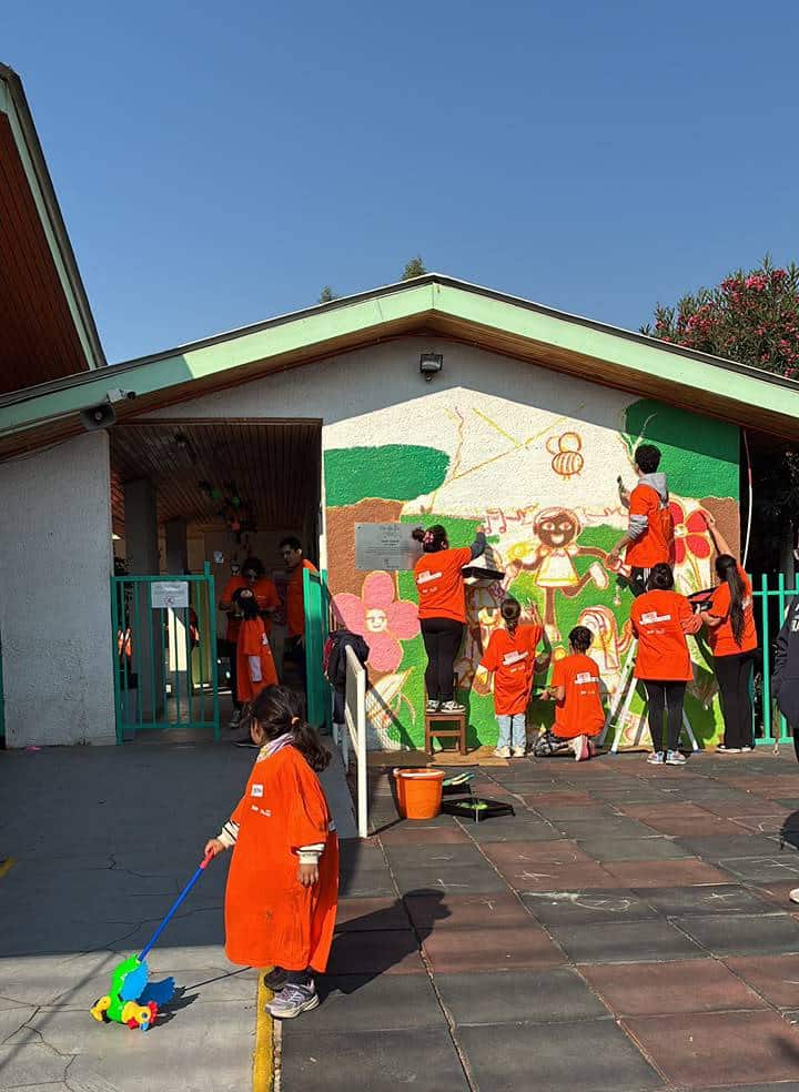En un día soleado, niños y adultos con camisetas naranjas colaboran en la pintura de un mural colorido en la pared exterior de un edificio educativo. Esta actividad comunitaria ilustra el esfuerzo colectivo por mejorar el entorno y crear un espacio acogedor y estimulante para una sala cuna universal, fomentando un ambiente propicio para el desarrollo de los niños. Un niño pequeño aparece en primer plano con un juguete de arrastre.