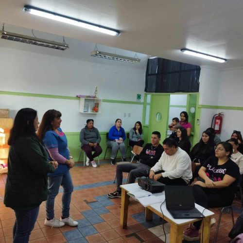 Reunión de apoderados o equipo en el Jardín Infantil Los Lagos. Dos educadoras de pie se dirigen a un grupo de adultos que las escuchan sentados en la sala.